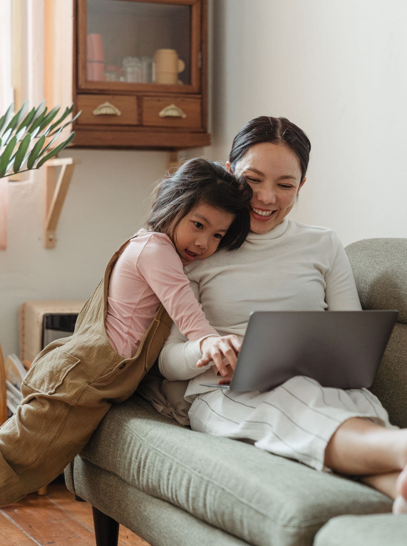 A mother and her young daughter sit close together on a sofa, smiling as they look at a laptop.