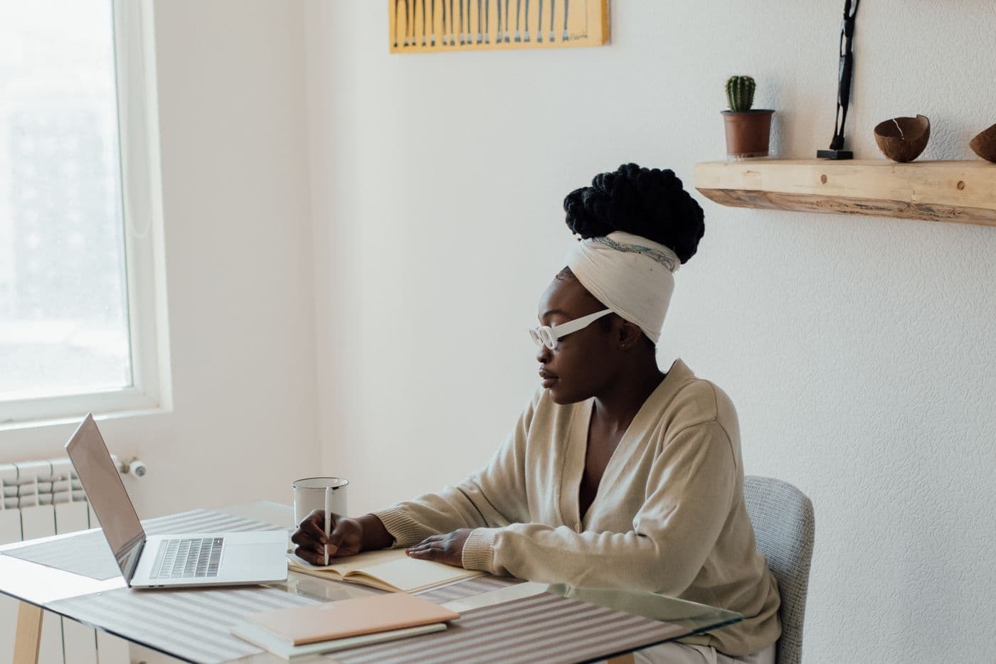 A woman working from home at a bright desk, writing notes beside her laptop.